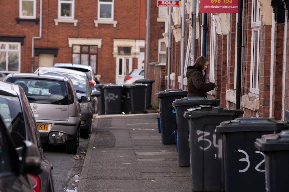A row of black wheeled rubbish bins with closed lids positioned along the pavement outside a red-brick residential building, with some bins featuring white graffiti markings. The bins are placed in front of windows with white frames, and the building's exterior includes decorative stone lintels above the windows. A woman wearing a dark brown coat is seen on the sidewalk, engaged in an activity near the bins, possibly preparing to dispose of waste or arrange the rubbish. Several parked cars, varying in color and model, are parked along the curb, partially visible on the left side of the image, with reflections on their windshields and windows indicating daylight. The environment suggests a typical urban street scene where private waste collection services, such as those provided by Rubbish Collection Hounslow, might be used for rubbish removal beyond local authority offerings. The scene is well-lit with natural light, emphasizing the textures of the brickwork, the plastic surfaces of the bins, and the overall tidy but utilitarian setting.