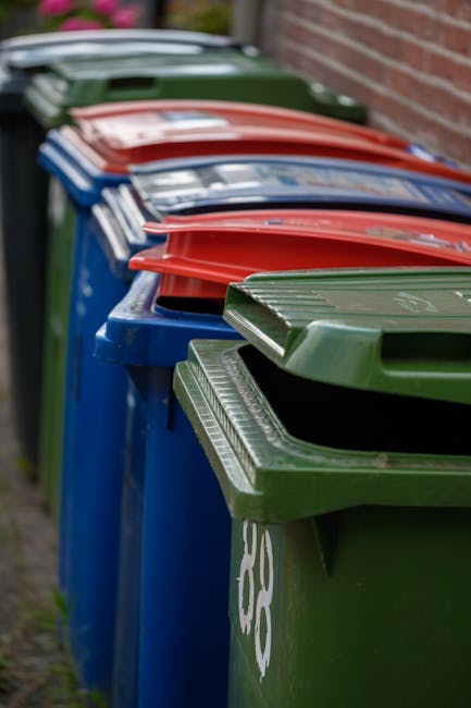 A row of large garden waste and rubbish bins positioned along a paved pathway beside a brick wall, with green and blue bins in the foreground, followed by red and other coloured lids further along. The bins are made of plastic with textured, ribbed lids, and are used for waste collection and disposal, indicating an outdoor setting possibly at a residential or estate location. The scene is illuminated by natural daylight, and the environment appears tidy, suggesting an organised approach to waste management and potential private rubbish collection services. The presence of multiple bins in a sequence aligns with a typical arrangement for waste collection, supporting services like those offered by Rubbish Collection Hounslow for bulky rubbish removal and estate clearance, particularly in areas like Cranford. The image emphasizes the importance of organised waste disposal solutions for residents and property managers engaged in alternative waste handling methods away from public authority collection.