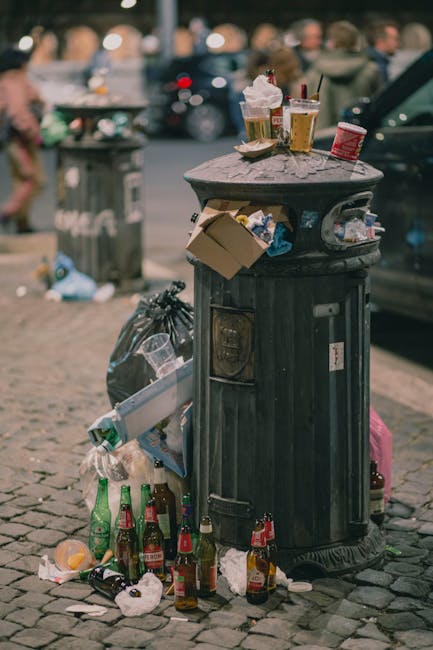 A black cylindrical public waste bin made of metal with a domed lid, positioned on a cobbled street. The bin is topped with scattered rubbish including plastic cups with remaining beverages, a crumpled paper bag, and a discarded cardboard box. The area around the base of the bin is cluttered with empty glass bottles, some with labels still attached, along with crumpled paper and other small debris. In the background, a slightly blurred street scene features parked cars, a grey trash receptacle, and pedestrians walking on the street. The overall scene suggests an outdoor urban environment where littering has occurred, highlighting the importance of effective rubbish collection services such as those provided by Rubbish Collection Hounslow for proper waste disposal and environmental cleanliness.