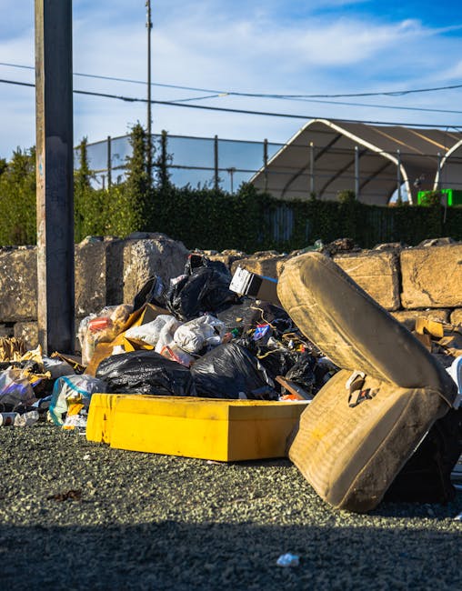 A pile of mixed household rubbish in an outdoor area on a gravel surface, including several black plastic garbage bags, a yellow plastic container, and a discarded car tyre made of rubber with visible tread and sidewall markings. The rubbish appears loosely arranged, with some bags torn open, revealing contents such as paper and plastic. In the background, there is a low stone wall constructed from irregularly shaped light-colored stones, behind which a dense hedge of bushes, a chain-link fence, and a solar panel-covered structure with curved metal frames are visible. Above, utility poles and overhead wires stretch across a partly cloudy sky with patches of blue. The scene suggests an instance of improper rubbish disposal, highlighting the need for professional waste collection services such as those offered by Rubbish Collection Hounslow, especially as part of alternative waste management outside of standard council collections.