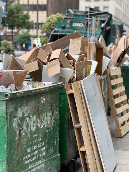A large green waste disposal bin filled to capacity with a mixture of cardboard boxes, some flattened and others partially assembled, along with white and brown packaging materials. The cardboard varies in size, color, and texture, with some showing printed labels or markings. Leaning against the bin on the right is a wooden pallet and a plastic-framed mirror with a silver reflective surface. In the background, a commercial garbage truck is partially visible with a blue-green body and hydraulic lifting mechanisms, positioned on a city street with multi-storey buildings and trees. The scene is outdoors under diffuse daylight, capturing the typical environment of private rubbish collection or on-site waste clearance handled by services such as Rubbish Collection Hounslow, serving the local Hounslow area.