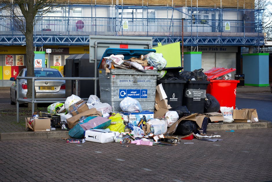 A person is seen disposing of paper waste into a grey rubbish bin with a black interior, located in an indoor environment. The individual is holding a crumpled brown paper bag in their right hand, positioning it above the open lid of the bin, while another similar paper bag is held in their left hand, slightly out of focus in the foreground. The bin is placed on a wooden surface, and behind it, part of a white background and some surrounding objects are visible, including a white cylindrical container on the right. The scene is well-lit with natural or artificial lighting, emphasizing the textures of the paper bags, which are rough and crinkled, contrasting with the smooth, glossy finish of the bin's exterior. This imagery reflects a typical waste disposal process suitable for private rubbish collection services like those offered by Rubbish Collection Hounslow, related to efficient on-site clearance of paper and light waste materials such as packaging or domestic trash, promoting organised rubbish management in residential or commercial settings.