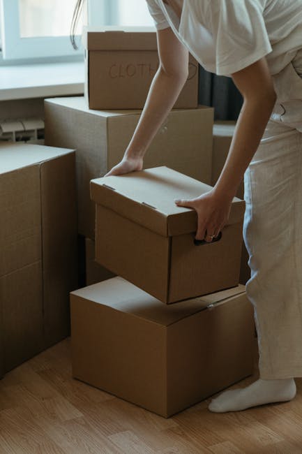 A person wearing white pants and a light-colored top is seen stacking and handling large cardboard boxes inside a room with natural light. The individual is placing a medium-sized, plain brown cardboard box on top of two other similar boxes, one of which has the word 'CLOTHES' handwritten in red marker on its lid. The boxes vary in size, with some stacked neatly and others arranged on a light wooden floor. In the background, additional boxes are visible, some sealed and others partially open, indicating an ongoing packing or clearing process. The room features a window with daylight streaming in, creating a soft, natural illumination across the scene. This environment suggests a domestic setting preparing for or facilitating private waste collection or move-related rubbish removal, aligning with services like those offered by Rubbish Collection Hounslow for on-site clearance and alternative waste handling.