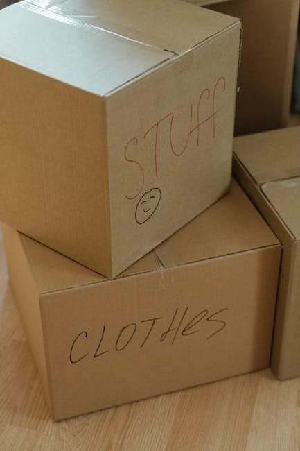 Two light brown cardboard boxes positioned on a wooden floor, with the larger box on top and a smaller one beneath. The larger box has handwritten text in red ink that reads 'STUFF' along with a small smiley face drawn below the text. The smaller box below is labeled in black ink with the word 'CLOTHES.' Both boxes appear to be sealed with tape and are situated indoors, likely in a storage or moving area. The scene suggests an organized approach to private waste handling or on-site clearance, typical of independent rubbish removal services such as those offered by Rubbish Collection Hounslow. The background shows part of another cardboard box, indicating a collection of packing or disposal materials prepared for collection or disposal, which aligns with typical domestic or commercial rubbish removal activities.