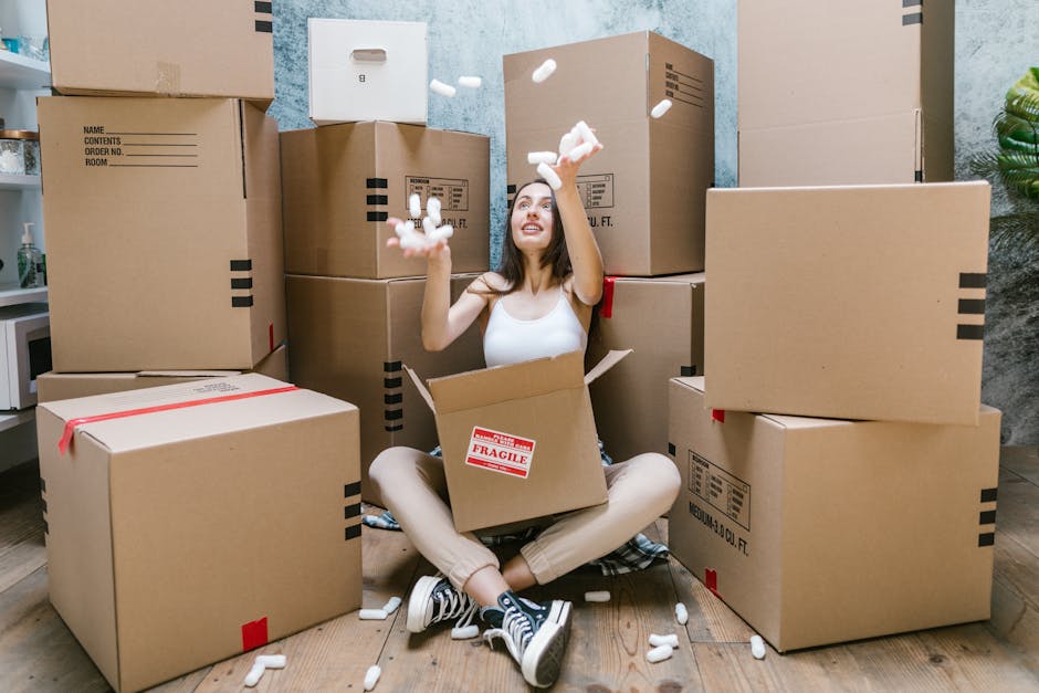 A person wearing white pants and a light-colored top is seen stacking and handling large cardboard boxes inside a room with natural light. The individual is placing a medium-sized, plain brown cardboard box on top of two other similar boxes, one of which has the word 'CLOTHES' handwritten in red marker on its lid. The boxes vary in size, with some stacked neatly and others arranged on a light wooden floor. In the background, additional boxes are visible, some sealed and others partially open, indicating an ongoing packing or clearing process. The room features a window with daylight streaming in, creating a soft, natural illumination across the scene. This environment suggests a domestic setting preparing for or facilitating private waste collection or move-related rubbish removal, aligning with services like those offered by Rubbish Collection Hounslow for on-site clearance and alternative waste handling.