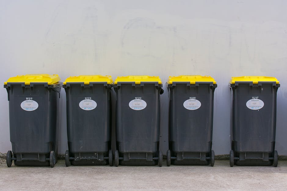 Five black wheeled trash bins with bright yellow lids are aligned side by side against a light gray wall, situated on a concrete surface. Each bin displays a white oval label with black text, and the bins are uniform in appearance, with some showing visible scuff marks or dirt on their surfaces. The yellow lids are securely closed, and the wheels are positioned at the bottom rear of each bin, with some showing slight wear. The environment is an outdoor or semi-outdoor setting, typical for waste collection points, with natural daylight illuminating the scene evenly. This arrangement suggests an organized private waste disposal or rubbish collection service such as Rubbish Collection Hounslow, which provides alternative waste handling options outside of municipal collection systems. The scene emphasizes the practicality of on-site rubbish storage for efficient waste management in residential or commercial properties, aligning with the context of rubbish removal and skip services often used for larger cleanups or garden waste collection, as referenced in the page about Osterley Park garden waste rubbish collection tips.