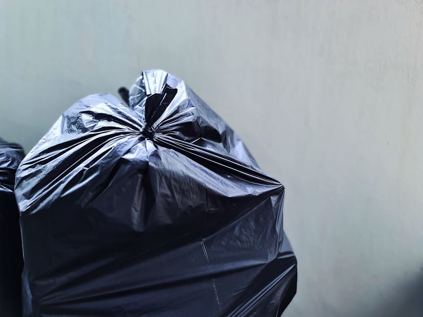 Close-up of a tightly tied black plastic rubbish bag placed against a light-colored, smooth wall. The bag appears to be filled with household waste and has a glossy, crinkled surface, showing creases and folds typical of plastic material. The top of the bag is gathered and secured with a knot, preventing spillage. In the background, additional black rubbish bags are partially visible, stacked or aligned on a surface, suggesting an outdoor or driveway setting prepared for collection. The lighting is neutral and evenly distributed, highlighting the sheen of the plastic and the folds of the bags. This image relates to private waste disposal and rubbish collection services, such as those offered by Rubbish Collection Hounslow, and exemplifies typical preparations for rubbish removal or on-site clearance, especially in the context of garden waste or household clutter suited for independent collection.