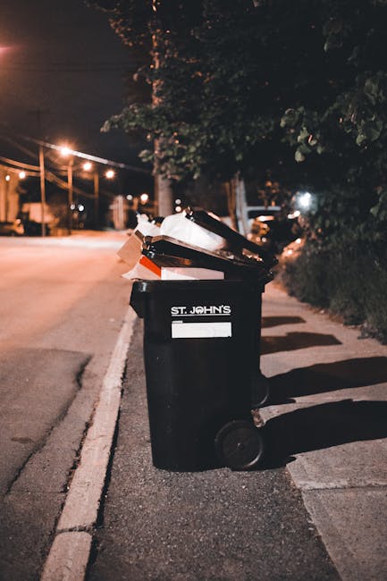 Five black wheeled trash bins with bright yellow lids are aligned side by side against a light gray wall, situated on a concrete surface. Each bin displays a white oval label with black text, and the bins are uniform in appearance, with some showing visible scuff marks or dirt on their surfaces. The yellow lids are securely closed, and the wheels are positioned at the bottom rear of each bin, with some showing slight wear. The environment is an outdoor or semi-outdoor setting, typical for waste collection points, with natural daylight illuminating the scene evenly. This arrangement suggests an organized private waste disposal or rubbish collection service such as Rubbish Collection Hounslow, which provides alternative waste handling options outside of municipal collection systems. The scene emphasizes the practicality of on-site rubbish storage for efficient waste management in residential or commercial properties, aligning with the context of rubbish removal and skip services often used for larger cleanups or garden waste collection, as referenced in the page about Osterley Park garden waste rubbish collection tips.