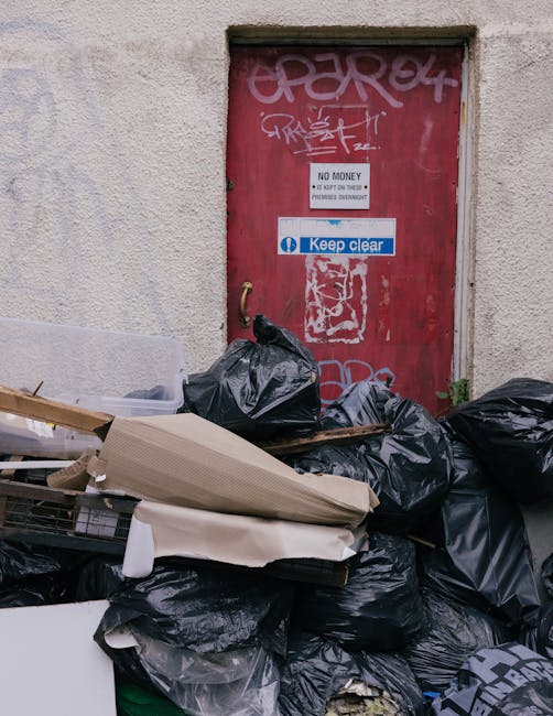 A red metal door with graffiti tags in white and pink spray paint is set into a rough-textured beige wall. A white sign with black text reading 'No Money' and additional smaller text is fixed above a blue and white 'Keep clear' notice attached directly below. In front of the door, there is a large pile of black garbage bags, some tied and others slightly open, revealing various waste materials. Among the bags, there are flattened cardboard boxes, including one large piece with layered cardboard edges, and a small plastic container. The waste is stacked on the paved ground, spilling out onto the surrounding pavement area, indicating a private rubbish collection or on-site clearance approaching or in progress. The scene suggests proximity to an alleyway or back entrance of a building, with environmental conditions under natural light, which illuminates the pile and highlights the industrial context of rubbish removal services.