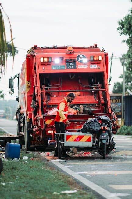 A waste collection worker wearing a yellow and red high-visibility vest is operating a large red refuse vehicle on the side of a paved street. The rear of the vehicle is open, revealing mechanical components and a loading area. The worker appears to be managing collected waste, which is contained in a black trash bag placed on a small metal platform attached to the vehicle. Nearby, there is a black motorcycle parked on the sidewalk, partly obscured by the waste truck. The surrounding environment includes a grassy verge with a small tree on the left, and in the background, there are commercial buildings and utility poles with overhead power lines. The scene is during daylight, with an overcast sky providing diffused lighting, capturing a typical urban rubbish collection process that could be associated with independent or private waste disposal services covered by rubbish removal regulations.